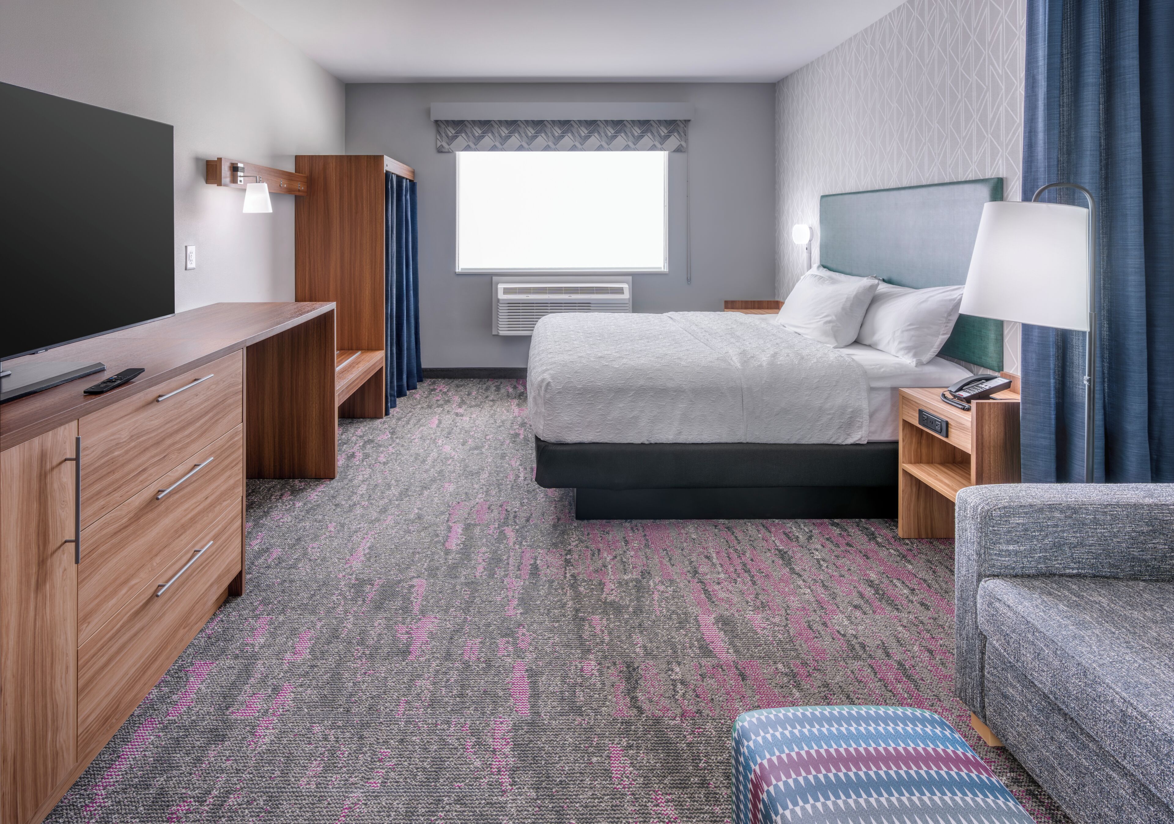 A hotel room with a bed, bedside table, lamp, TV, dresser, and a window with curtains.