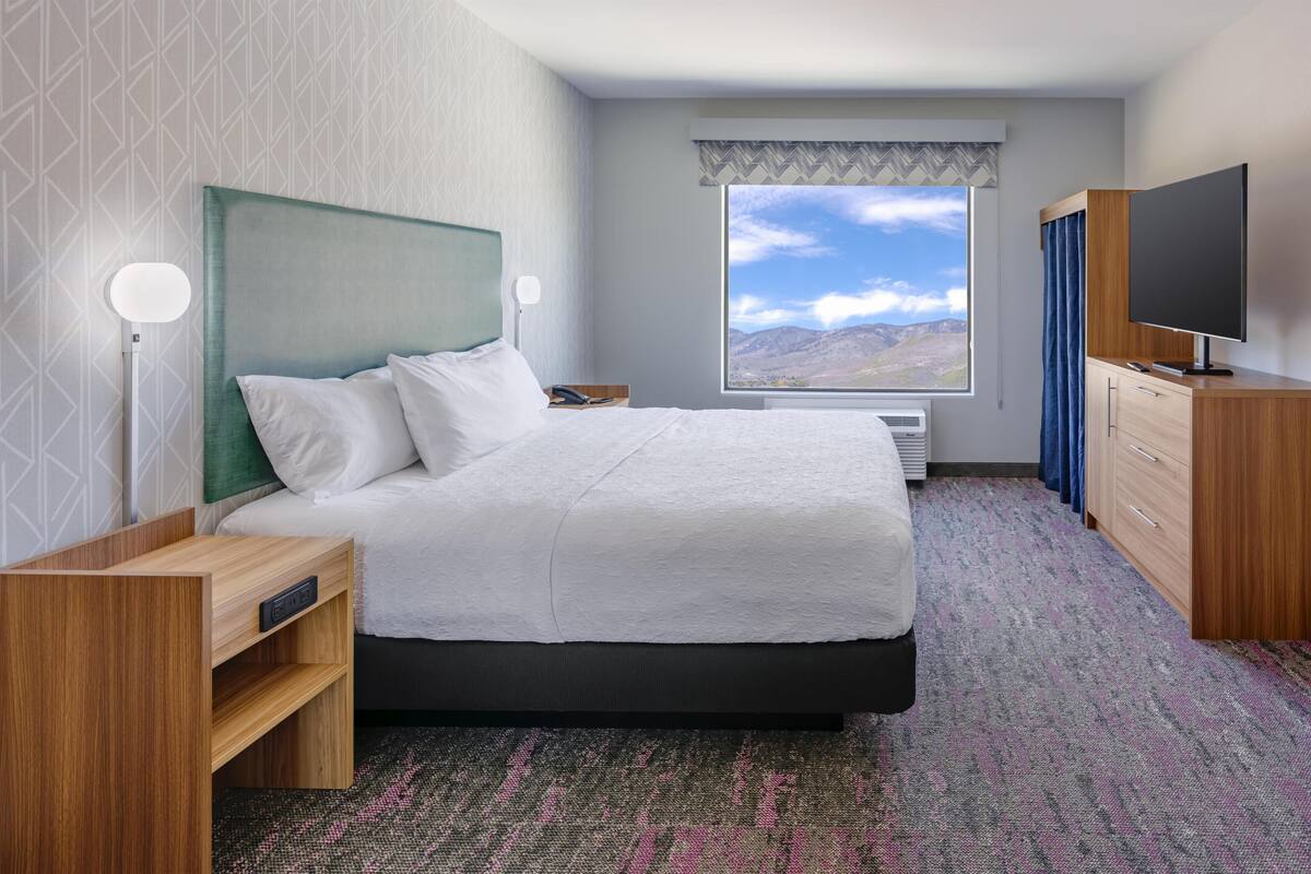 A hotel room with a bed, bedside table, TV, wardrobe, and a window with a mountain view.