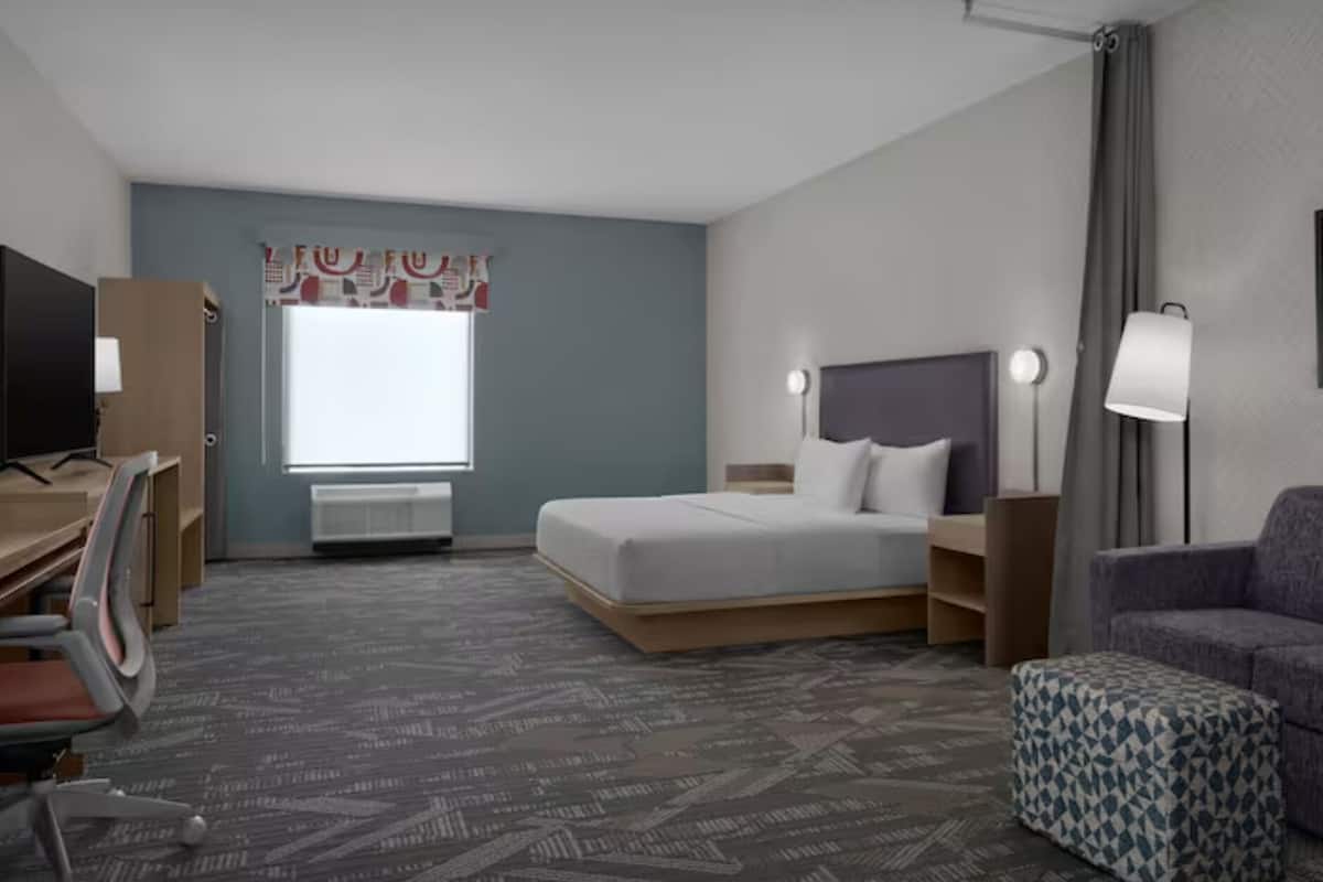 Una habitación de hotel con cama, escritorio, silla, sofá y ventana con cortinas.