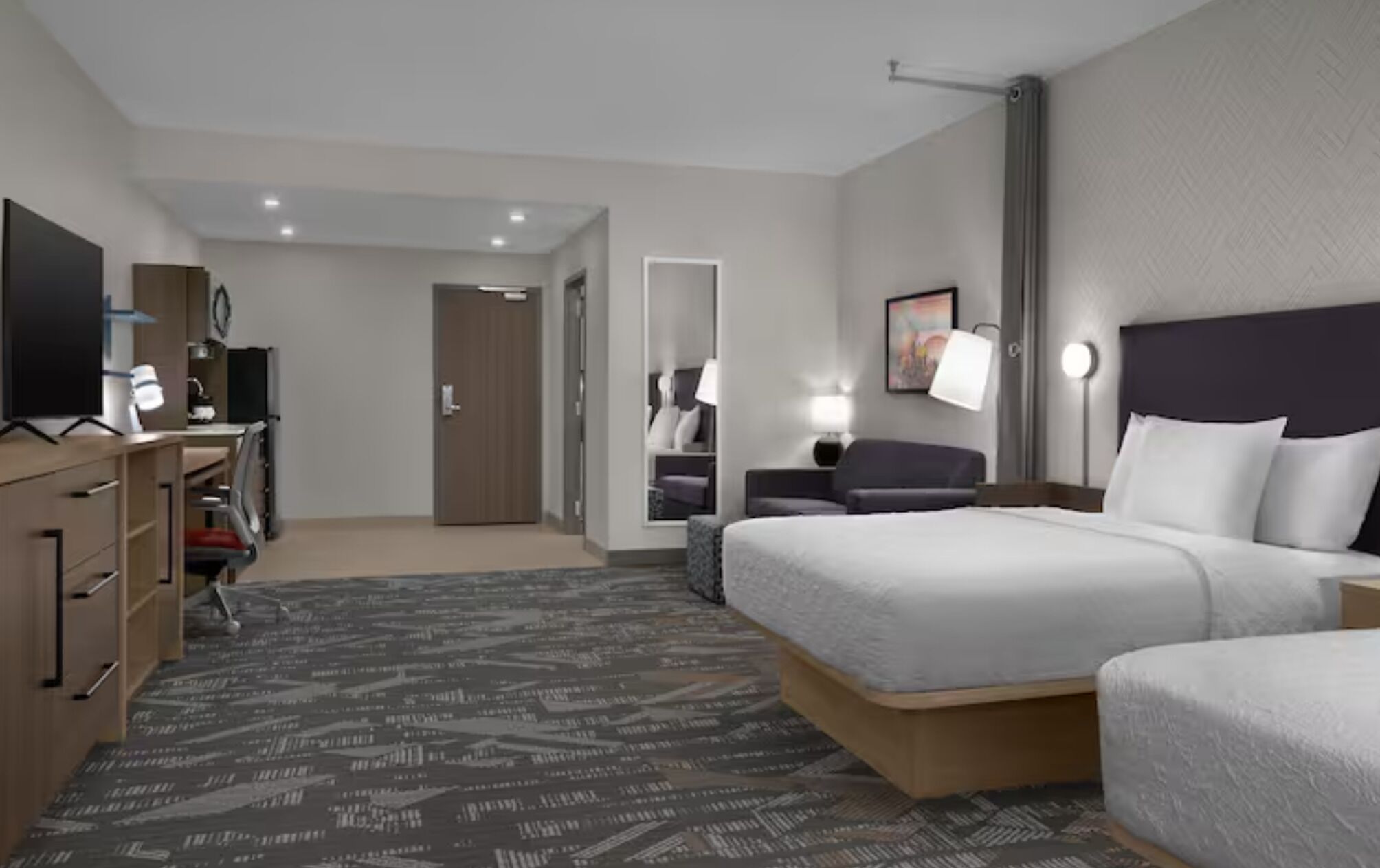A hotel room with a bed, a desk with a computer, a dresser, a mirror, and a door.