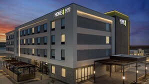 A modern hotel with 'HOME 2 SUITES BY HILTON' signage, a covered outdoor seating area, and a well-lit entrance.