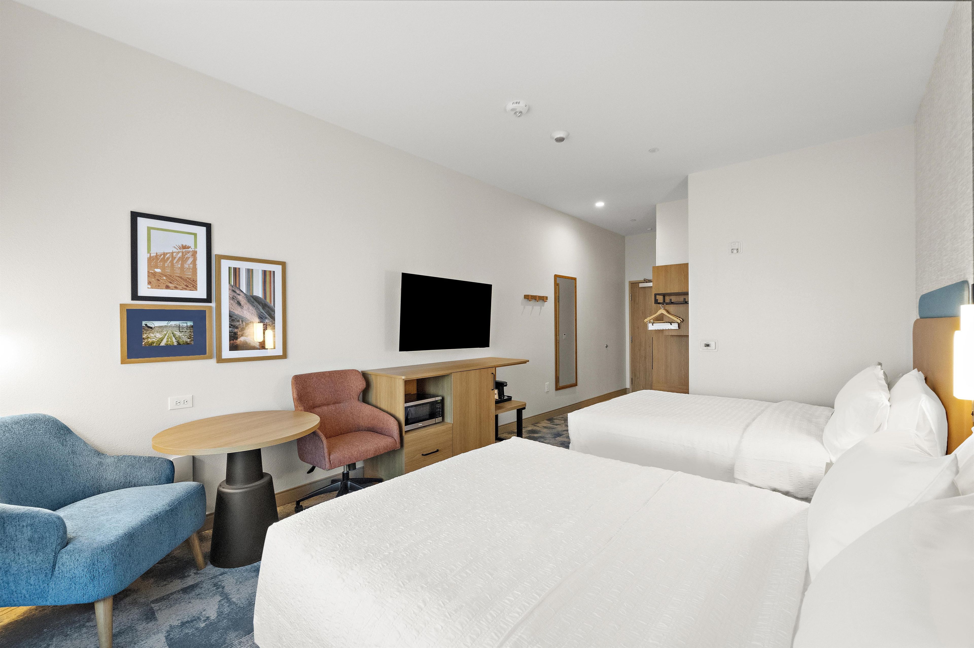 Une chambre d’hôtel avec deux lits, un bureau doté d’un téléviseur à écran plat, une chaise, une petite table et un téléviseur fixé au mur.
