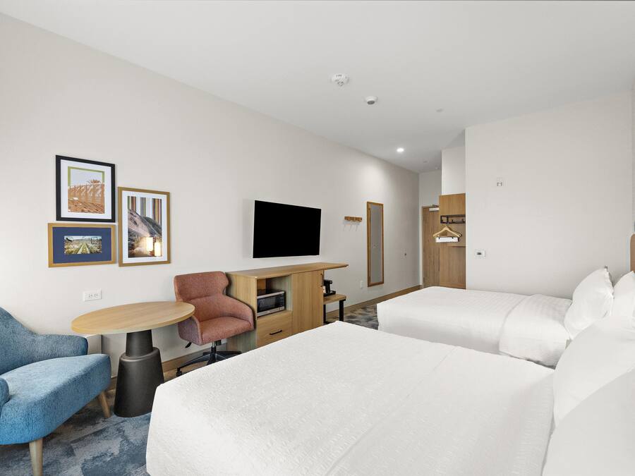 Une chambre d’hôtel avec deux lits, un bureau doté d’un téléviseur à écran plat, une chaise, une petite table et un téléviseur fixé au mur.