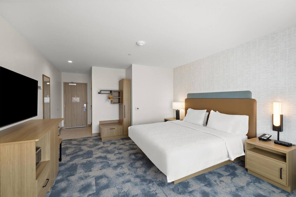 Een hotelkamer met een bed, nachtkastjes, een televisie en een deur naar een gang.