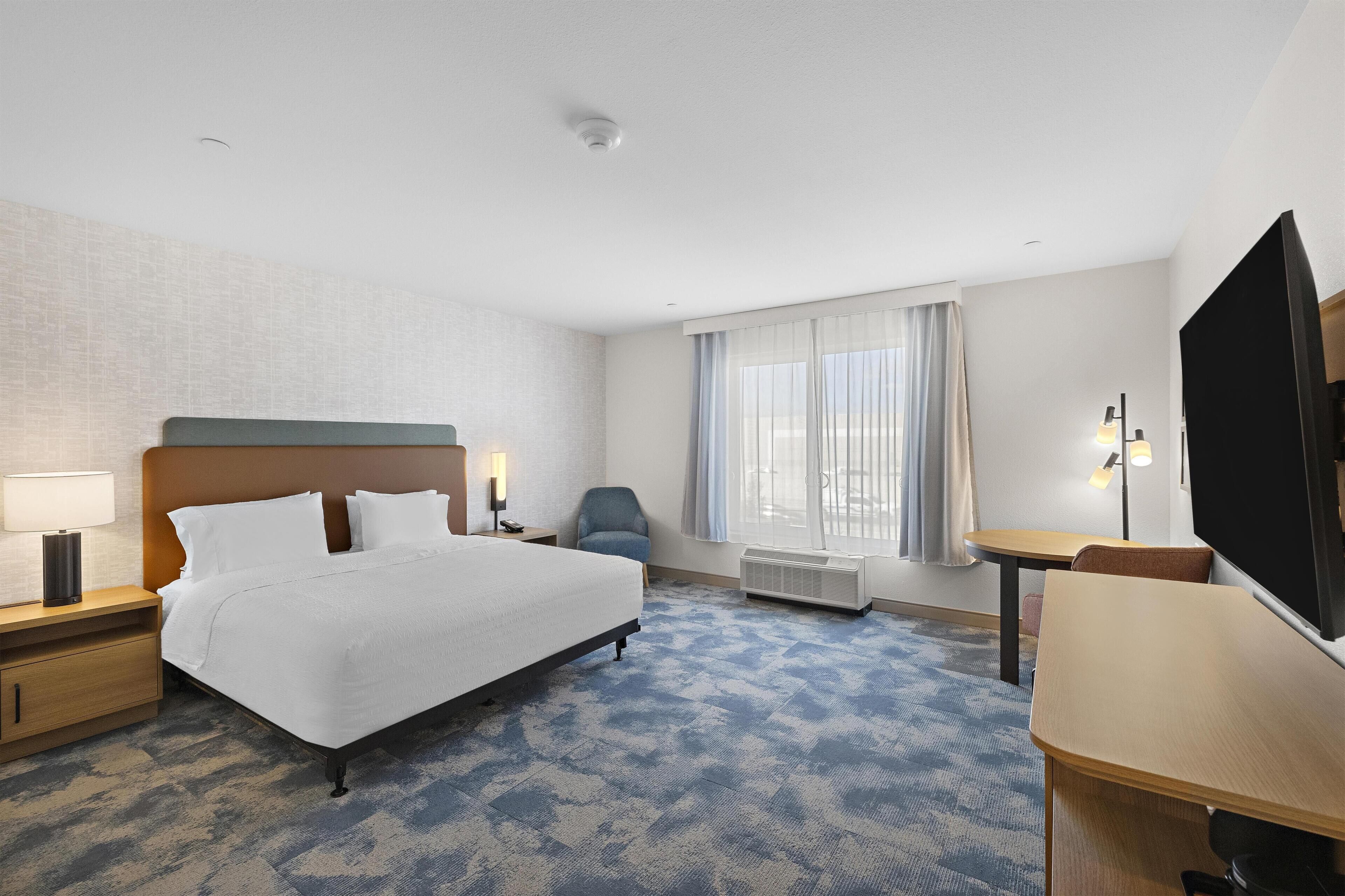 Una habitación de hotel con cama, escritorio, silla, televisor y ventana con cortinas.