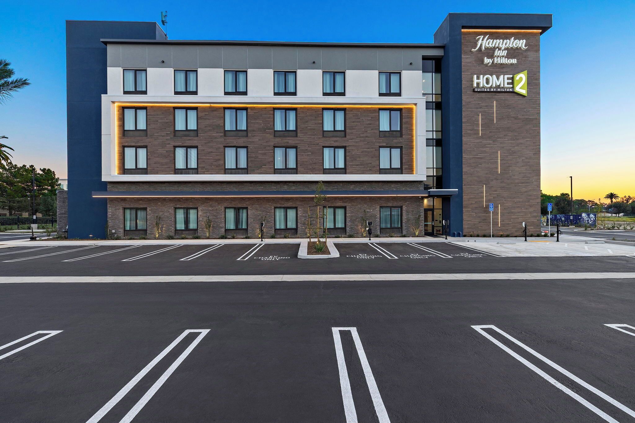 A modern hotel building with multiple floors, a prominent sign for Hampton Inn by Hilton HOME 2, and a parking lot with marked spaces.