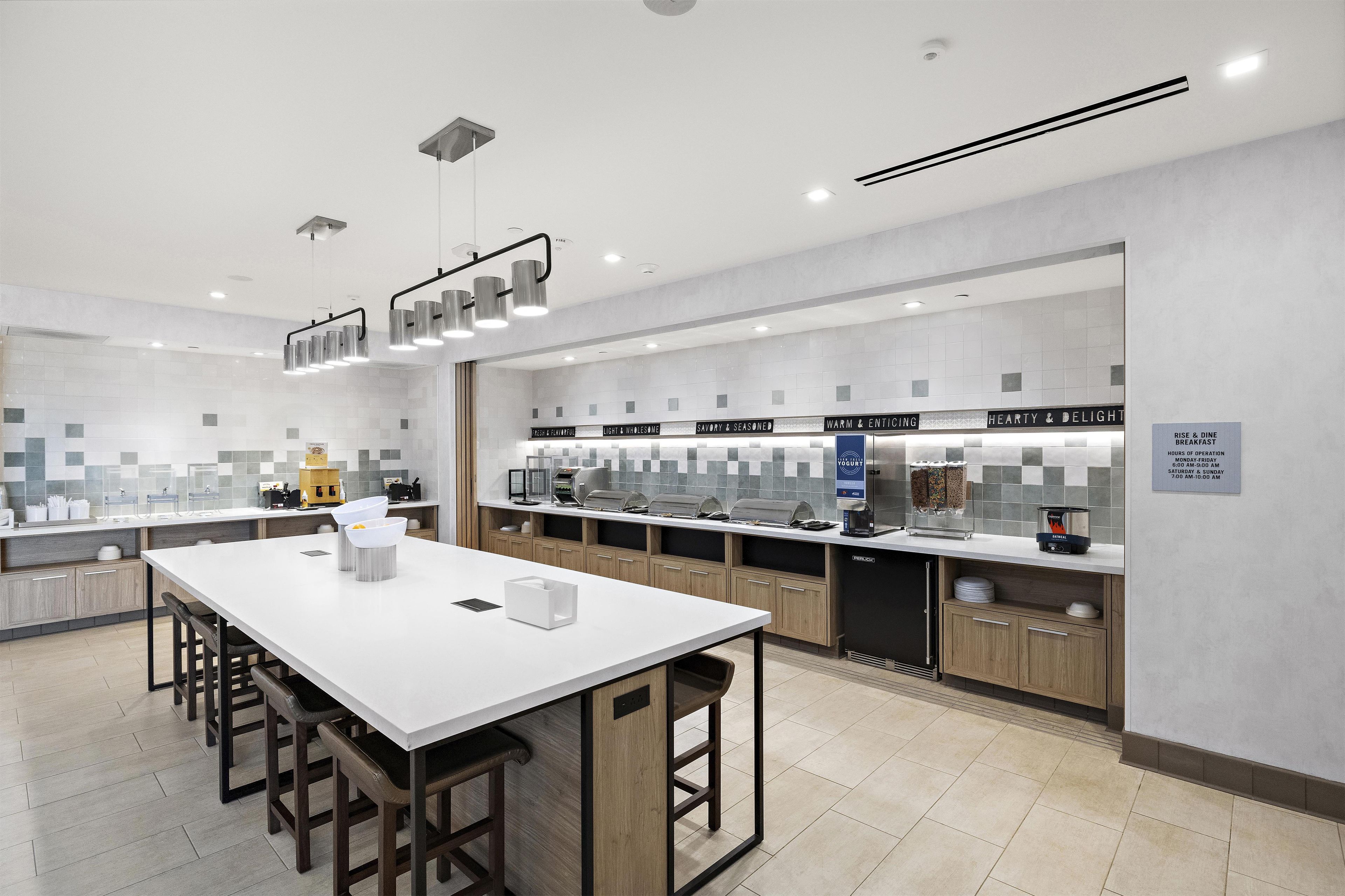 Une cuisine moderne dotée d’un îlot central, de tabourets de bar, d’un coin café et d’un panneau indiquant les heures du petit-déjeuner.