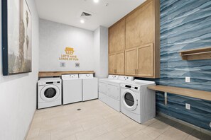 Une buanderie avec quatre machines à laver blanches, des armoires en bois, un mur bleu à texture et un panneau indiquant : « AMUSONS-NOUS À FOND ».