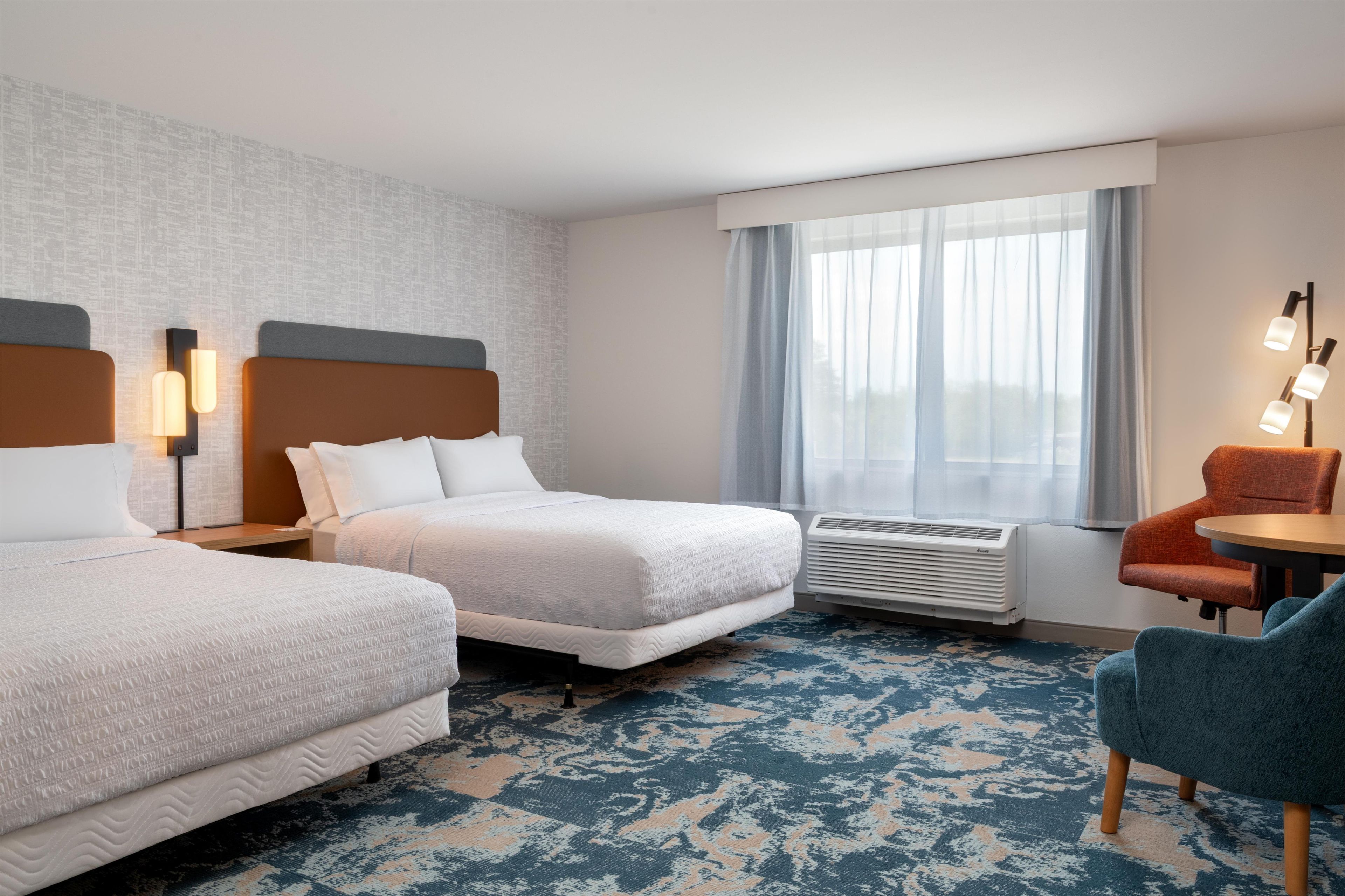 A hotel room with two beds, a desk, a chair, a window with curtains, and an air conditioning unit.