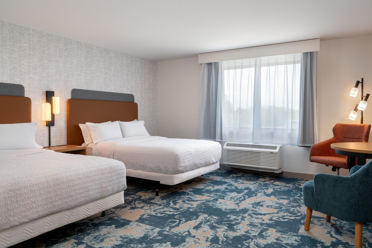 A hotel room with two beds, a desk, a chair, a window with curtains, and an air conditioning unit.