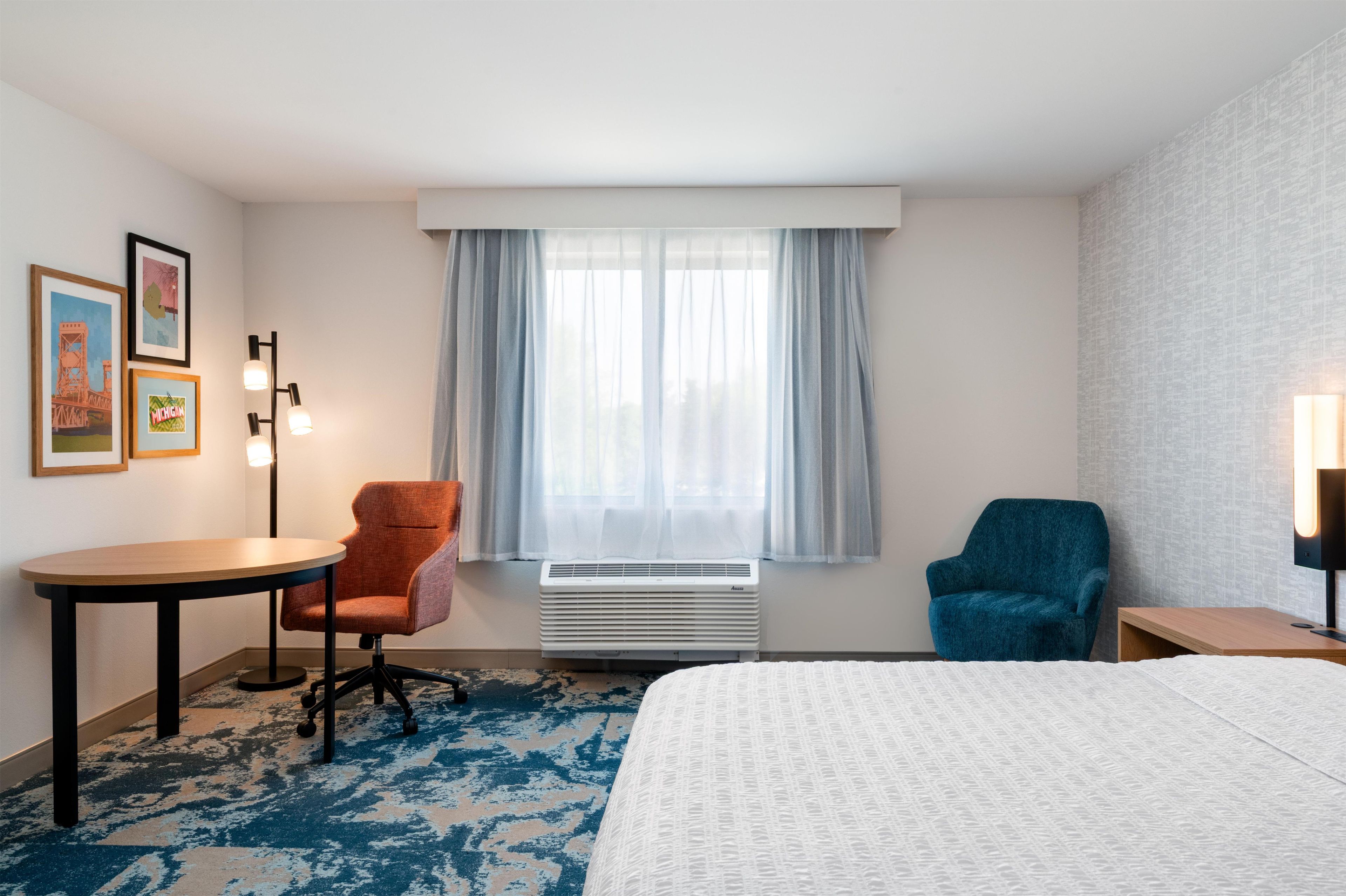 A hotel room with a bed, a desk, two chairs, a window with curtains, and wall-mounted lights.