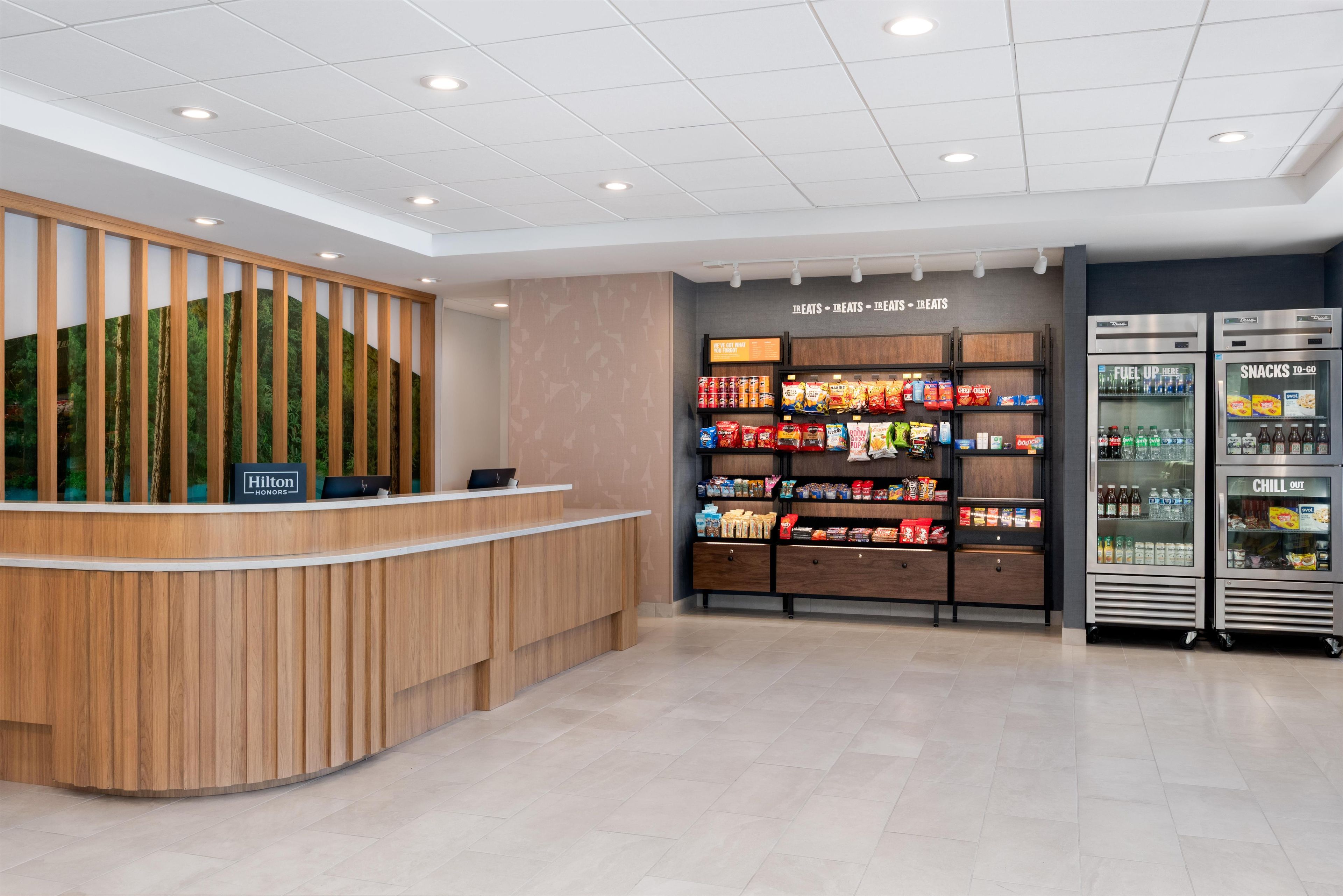 Zona de recepción de un hotel con un mostrador de madera, una pantalla mural que muestra el logotipo de Hilton Honors y una máquina expendedora de aperitivos con la etiqueta 'SNACKS TO-GO'.