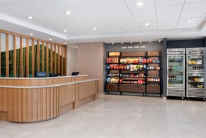 Uma área de recepção de hotel com um balcão de madeira, uma tela na parede exibindo o logotipo Hilton Honors e uma máquina de venda automática de lanches com a etiqueta 'LANCHES PARA LEVAR'.