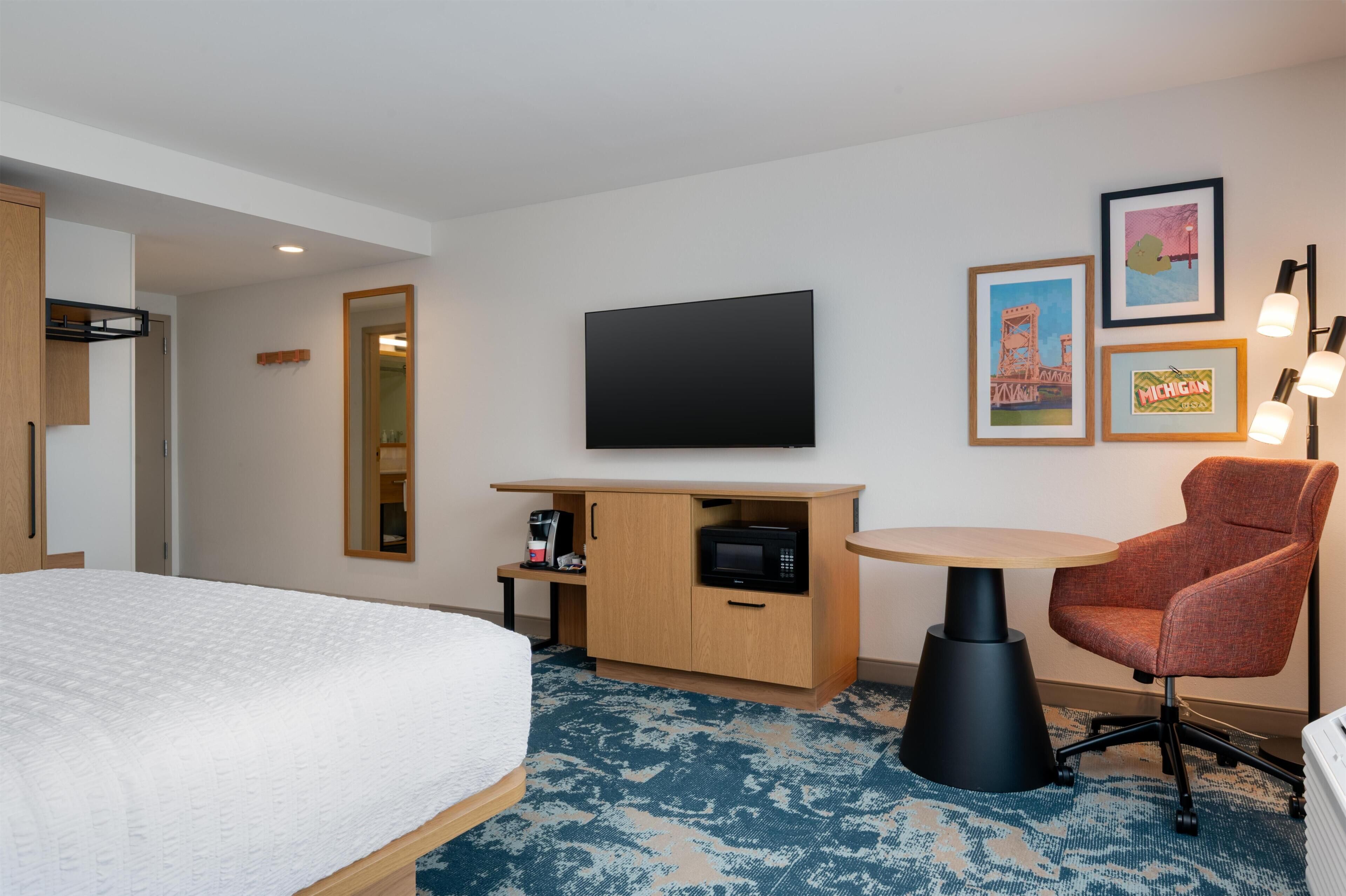 Habitación de hotel con cama, escritorio, televisor de pantalla plana, microondas, silla y mesita.