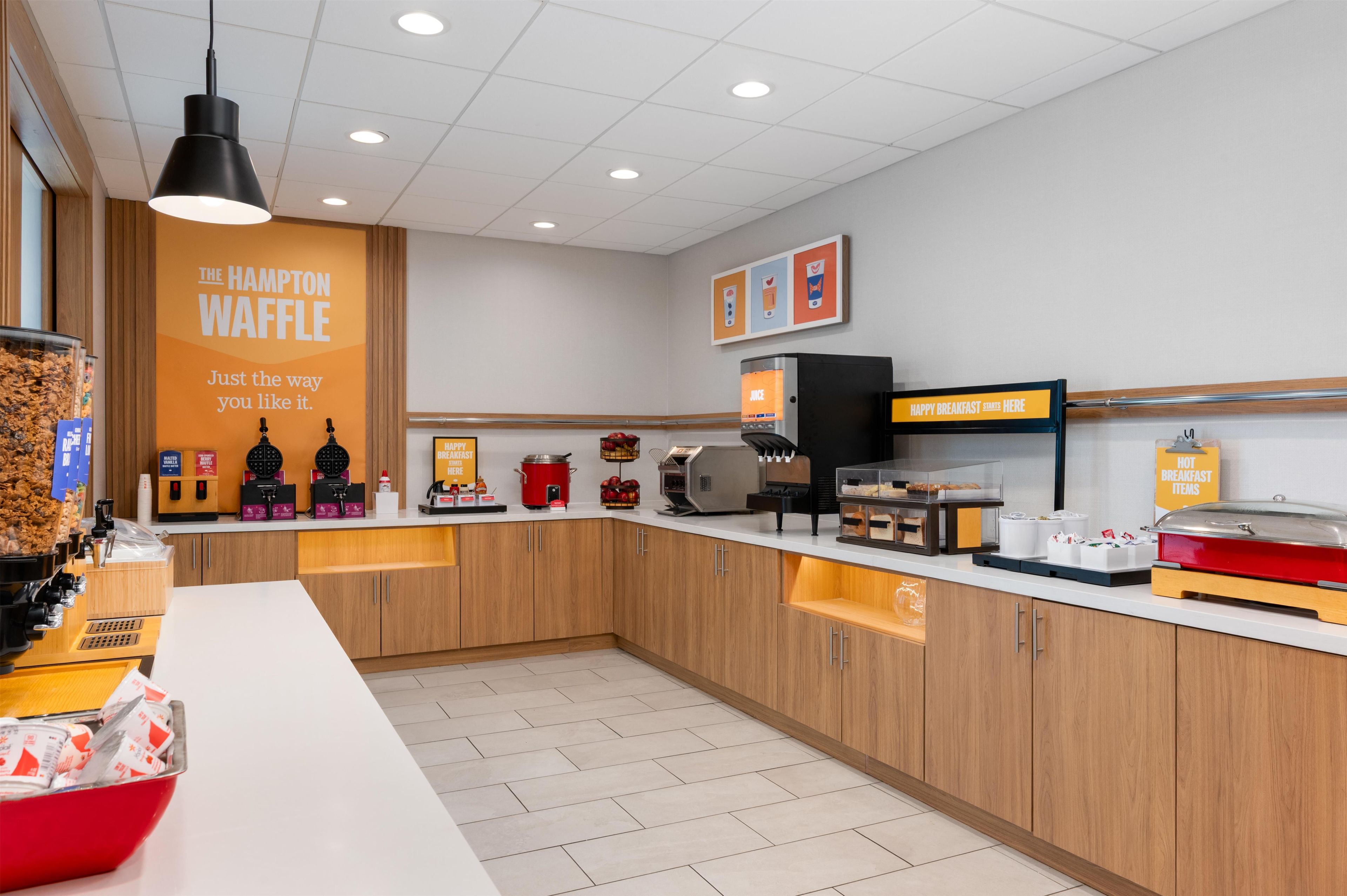 Un área de desayuno con máquinas para hacer gofres, dispensadores de jugo y un letrero que dice 'The Hampton Waffle'.