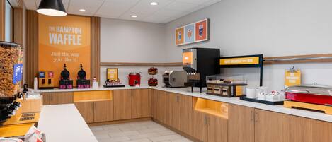 A breakfast area with waffle makers, juice dispensers, and a sign for 'The Hampton Waffle'.