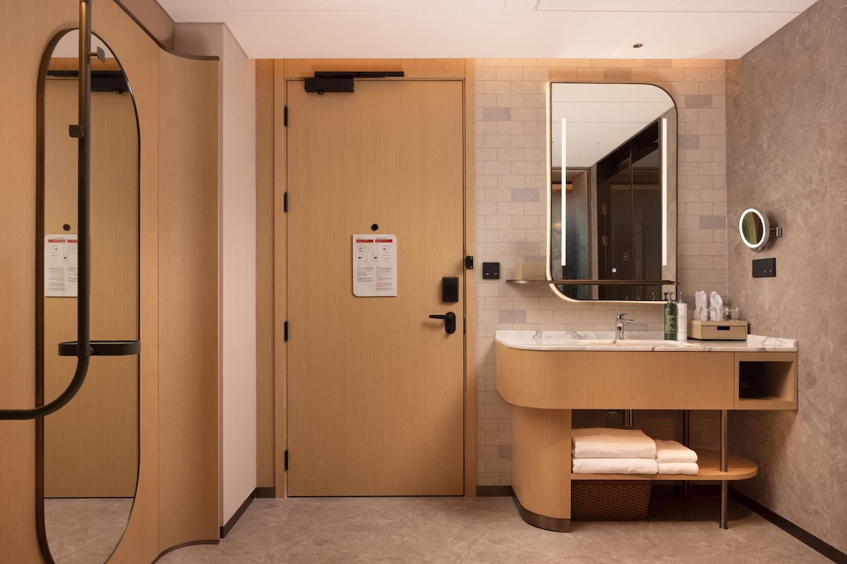 A modern bathroom with a wooden door, a large mirror, a sink with a marble countertop, and neatly folded towels.