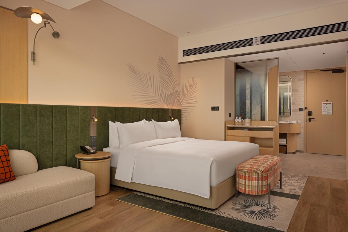 A hotel room with a bed, a bedside table with a lamp, a small sofa, a mirror, and a bathroom visible through an open door.