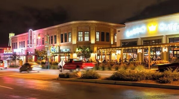 A street view at night with illuminated buildings, including a restaurant with outdoor seating and a brightly lit sign.