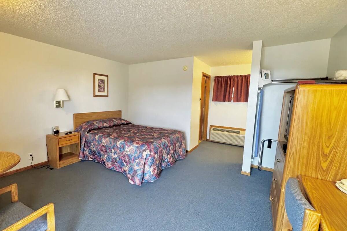 A small, neatly arranged room with a bed, a desk, a chair, a lamp, and a refrigerator.