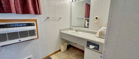 Un baño con tocador blanco, espejo con iluminación, calefactor y cubo de basura.
