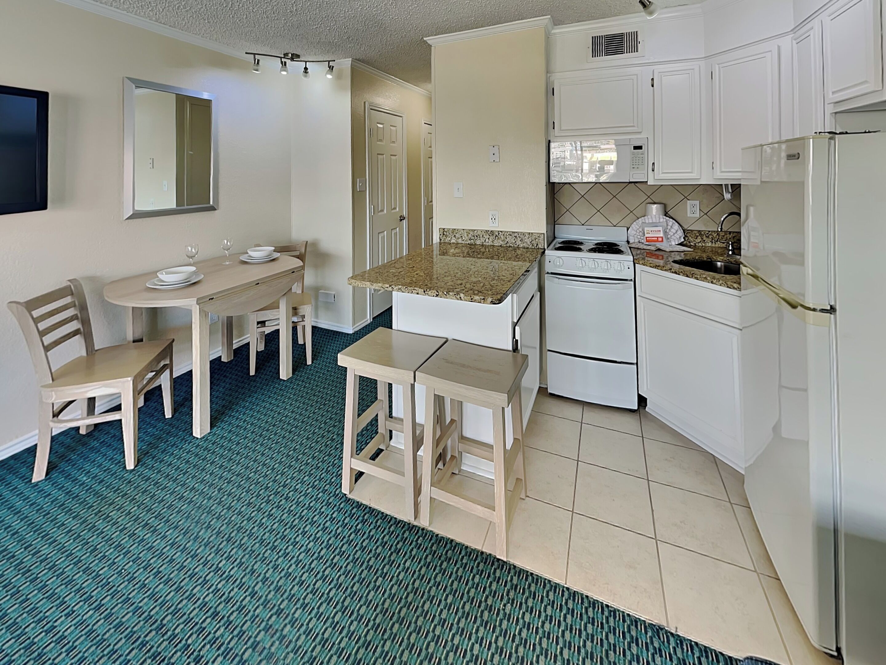 Une cuisine compacte avec des meubles blancs, un four à micro-ondes, une cuisinière et un réfrigérateur. On y trouve une table à manger avec des chaises et un petit tabouret.