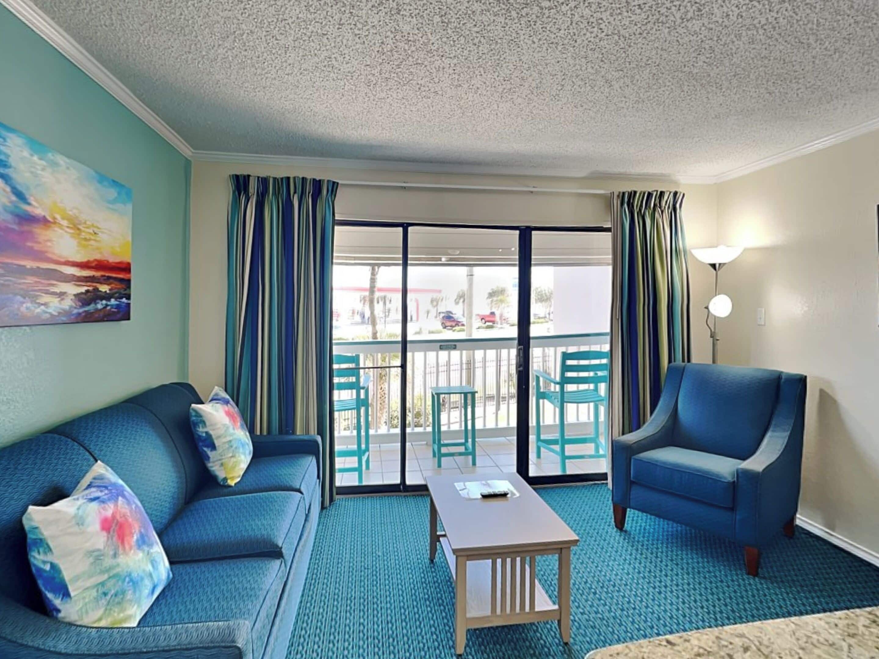 A hotel room with a blue sofa, a matching armchair, a small table, and a view of the outside through a sliding glass door.