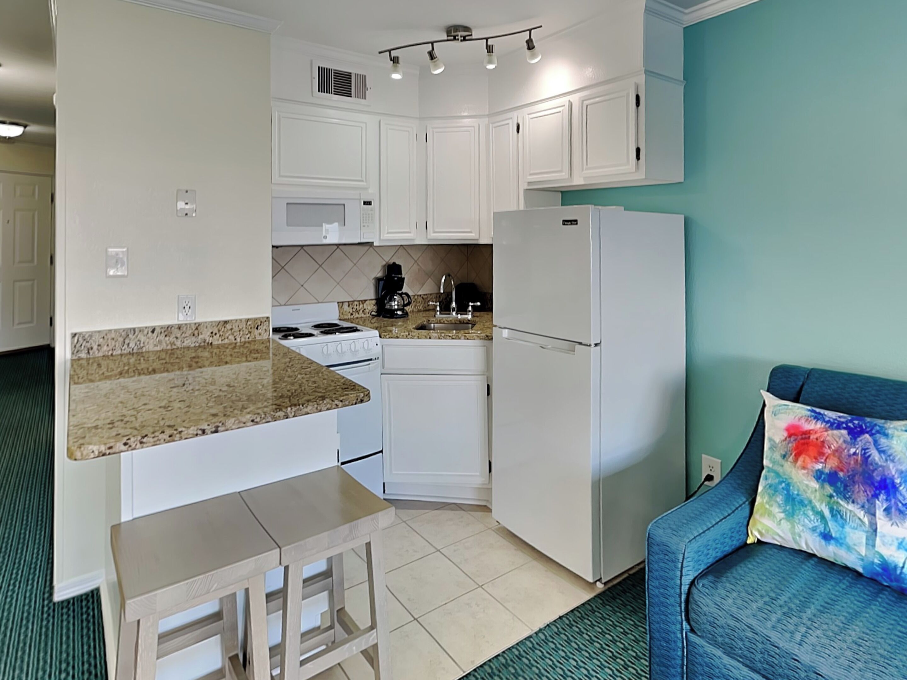 A compact kitchen with white cabinets, a stove, a refrigerator, and a microwave. There's a small wooden table and a blue chair with a colorful pillow.