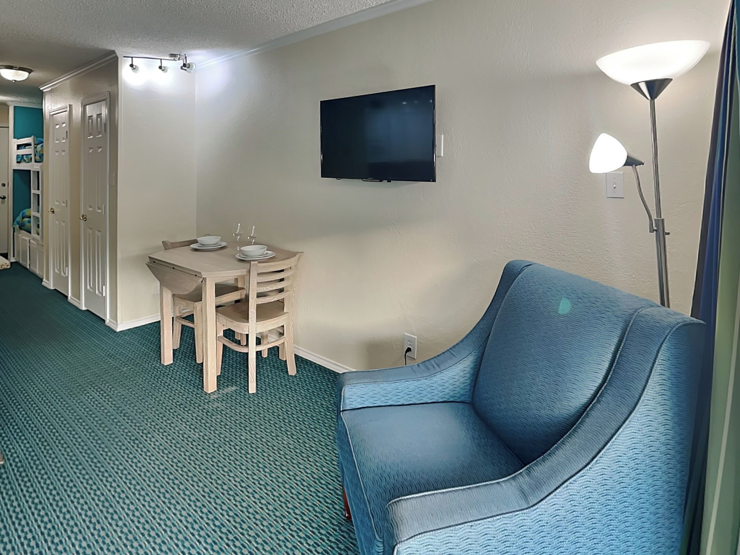 Een hotelkamer met een eettafel, stoelen, een aan de muur bevestigde televisie, een blauwe fauteuil en een vloerlamp.
