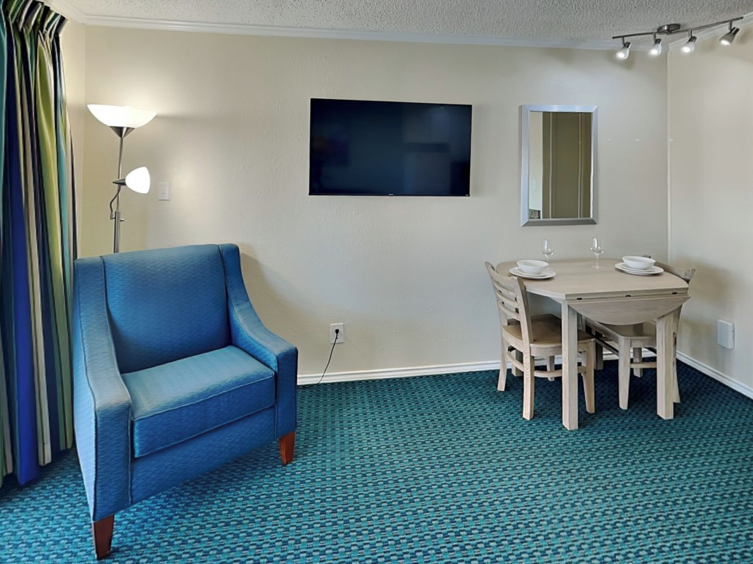 A hotel room with a blue armchair, a dining table with chairs, a flat-screen TV mounted on the wall, and a mirror above the table.