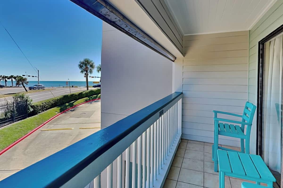 A balcony with a view of the street and ocean, two turquoise chairs, and a white railing.