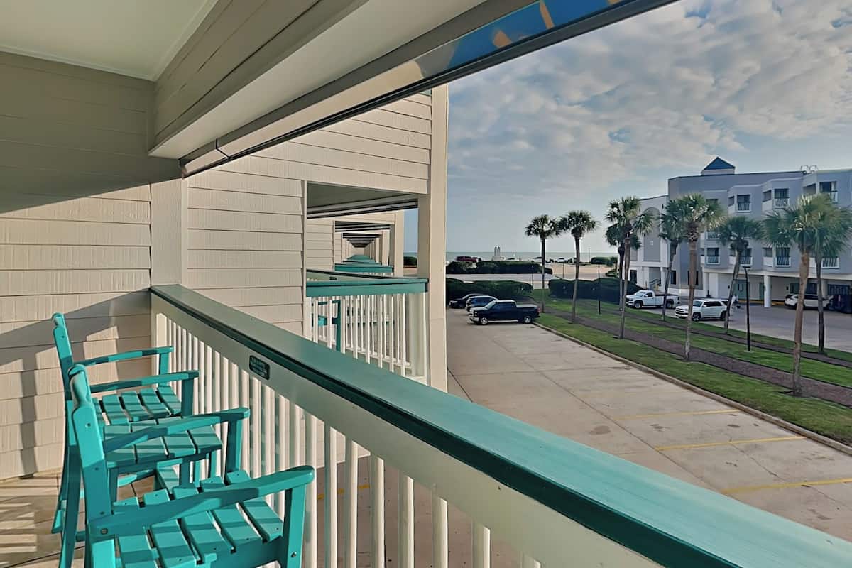 A balcony with teal chairs and a view of a parking lot and buildings.