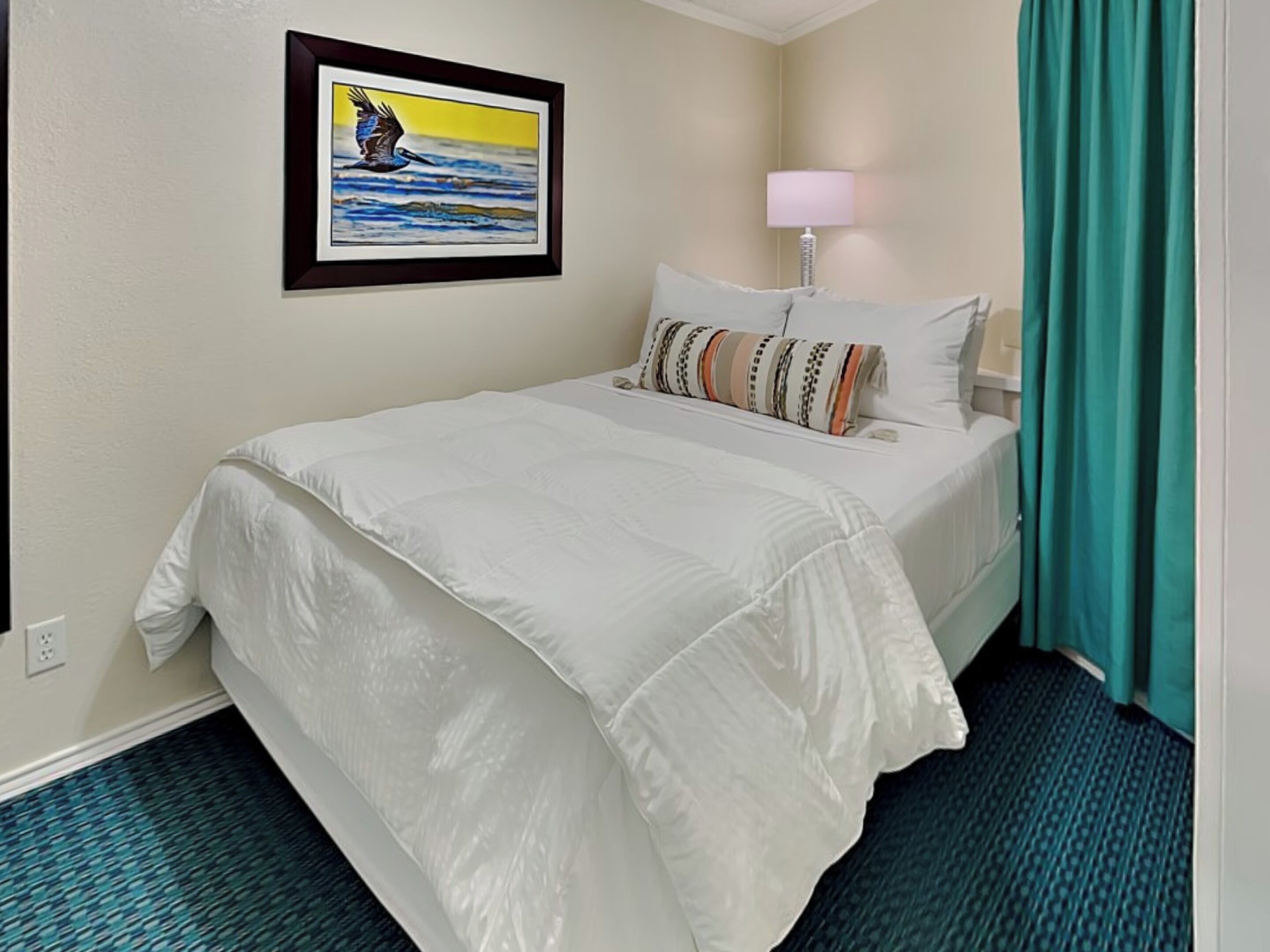 A hotel room with a bed, a framed picture on the wall, a lamp, and a teal curtain.