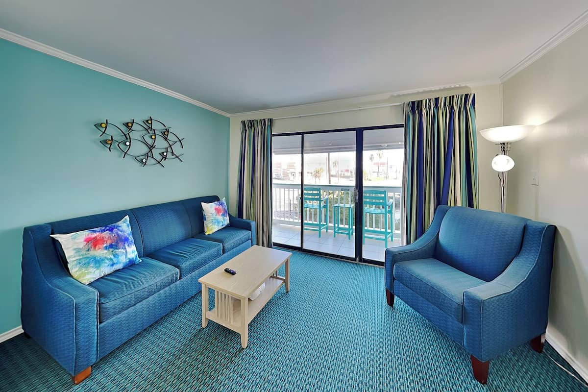 A living room with a blue sofa, a matching armchair, a small wooden table, and a view of the outside balcony through large glass doors.
