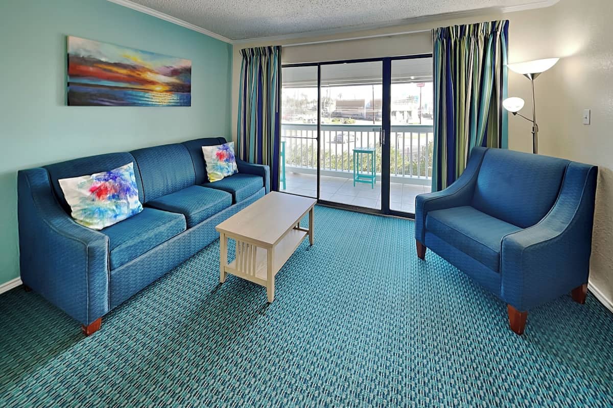 A room with a blue sofa, a matching armchair, a wooden coffee table, and a view through a large sliding glass door to an outdoor balcony.