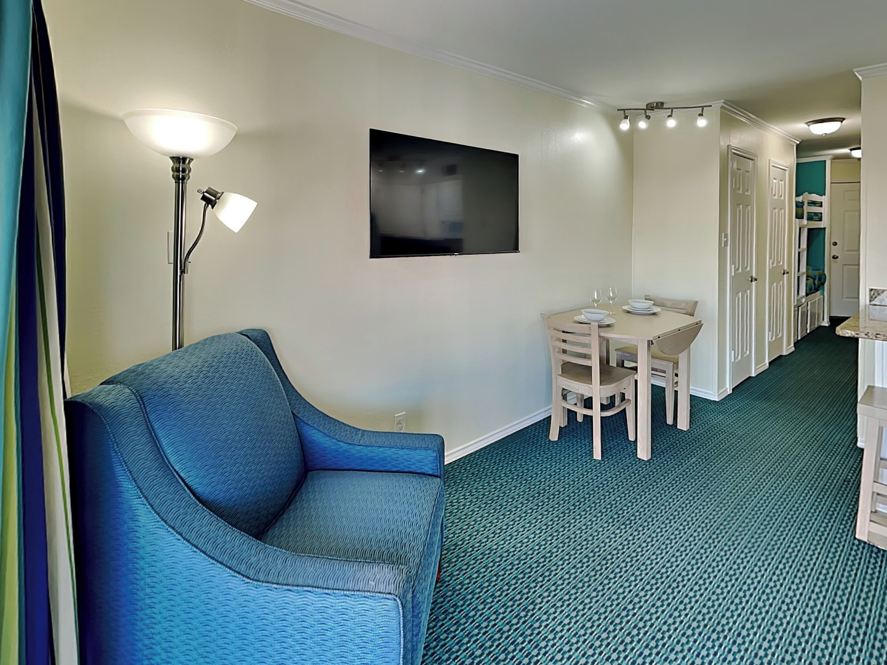 Une chambre d’hôtel avec un canapé bleu, une table à manger avec des chaises, un téléviseur à écran plat et un couloir avec des portes.