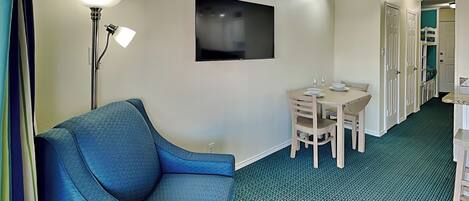 Une chambre d’hôtel avec un canapé bleu, une table à manger avec des chaises, un téléviseur à écran plat et un couloir avec des portes.