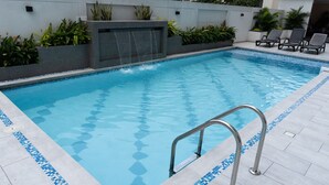 A rectangular swimming pool with a waterfall feature, surrounded by a tiled deck and plants.