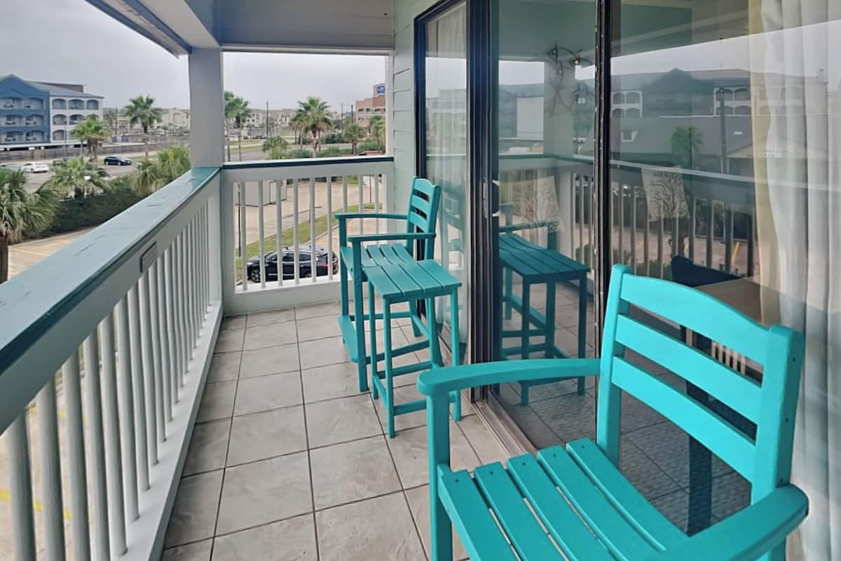 A balcony with turquoise chairs and a table, overlooking a street with palm trees and buildings.