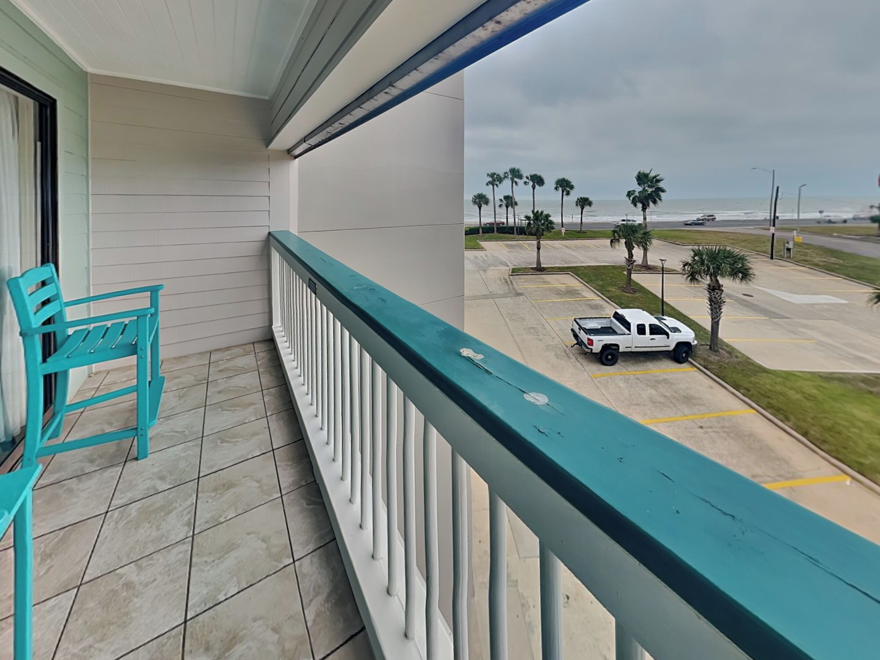 A balcony with a turquoise railing overlooking a parking area with palm trees and a view of the ocean.