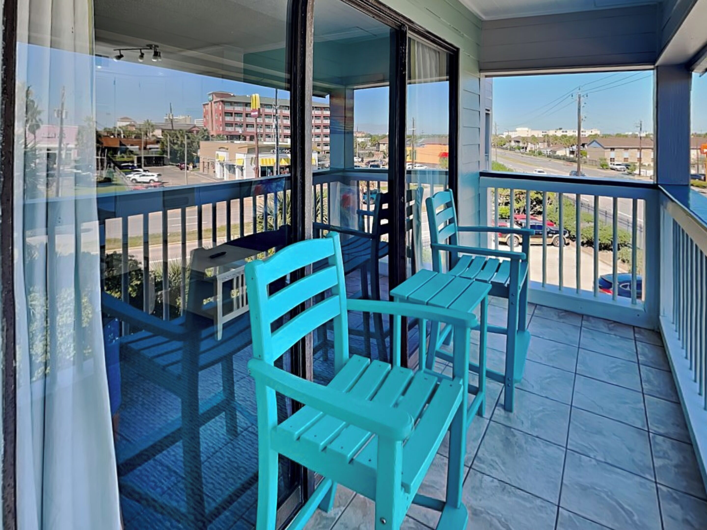 Un balcon avec deux chaises turquoise et une petite table, donnant sur une rue bordée d’immeubles et de voitures garées.