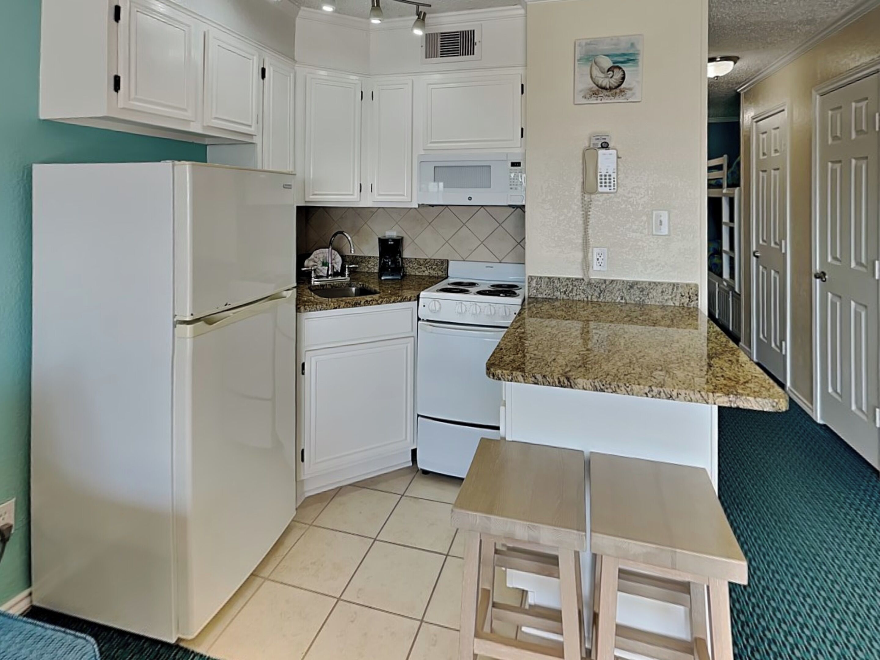 A compact kitchen with white cabinets, a white refrigerator, a stove, and a coffee maker. There is a small wooden table and a phone on the wall.