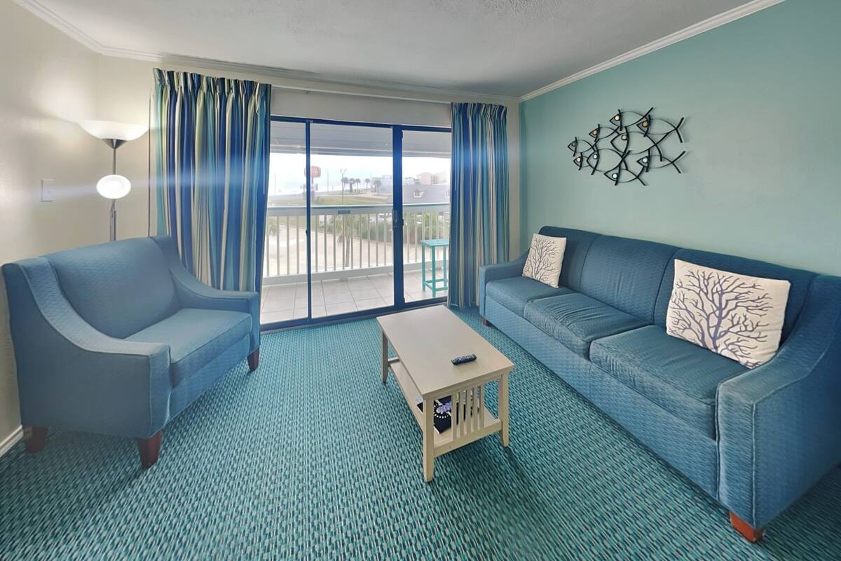 A room with a blue sofa, a blue armchair, and a small wooden coffee table. There is a large window with curtains, a wall-mounted light fixture, and a decorative wall piece.