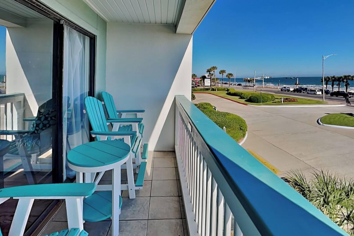 A balcony with turquoise chairs and a glass table, overlooking a coastal road with palm trees and a clear blue sky.