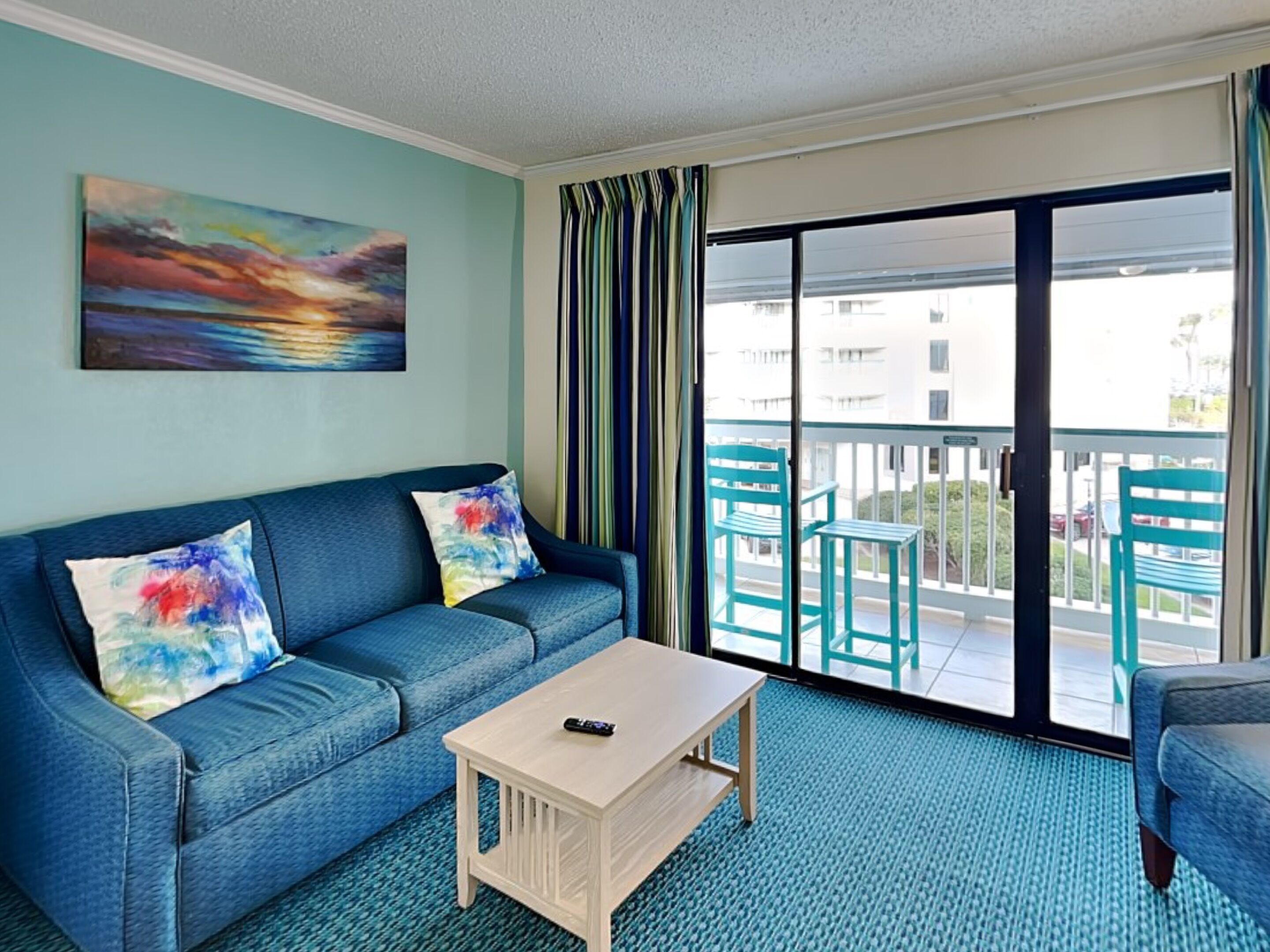 A room with a blue sofa, a small wooden table, and a balcony with chairs.