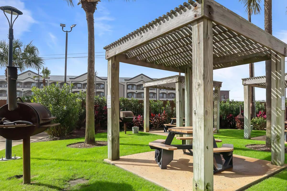 A wooden gazebo with a pergola roof, surrounded by a well-maintained lawn and palm trees.
