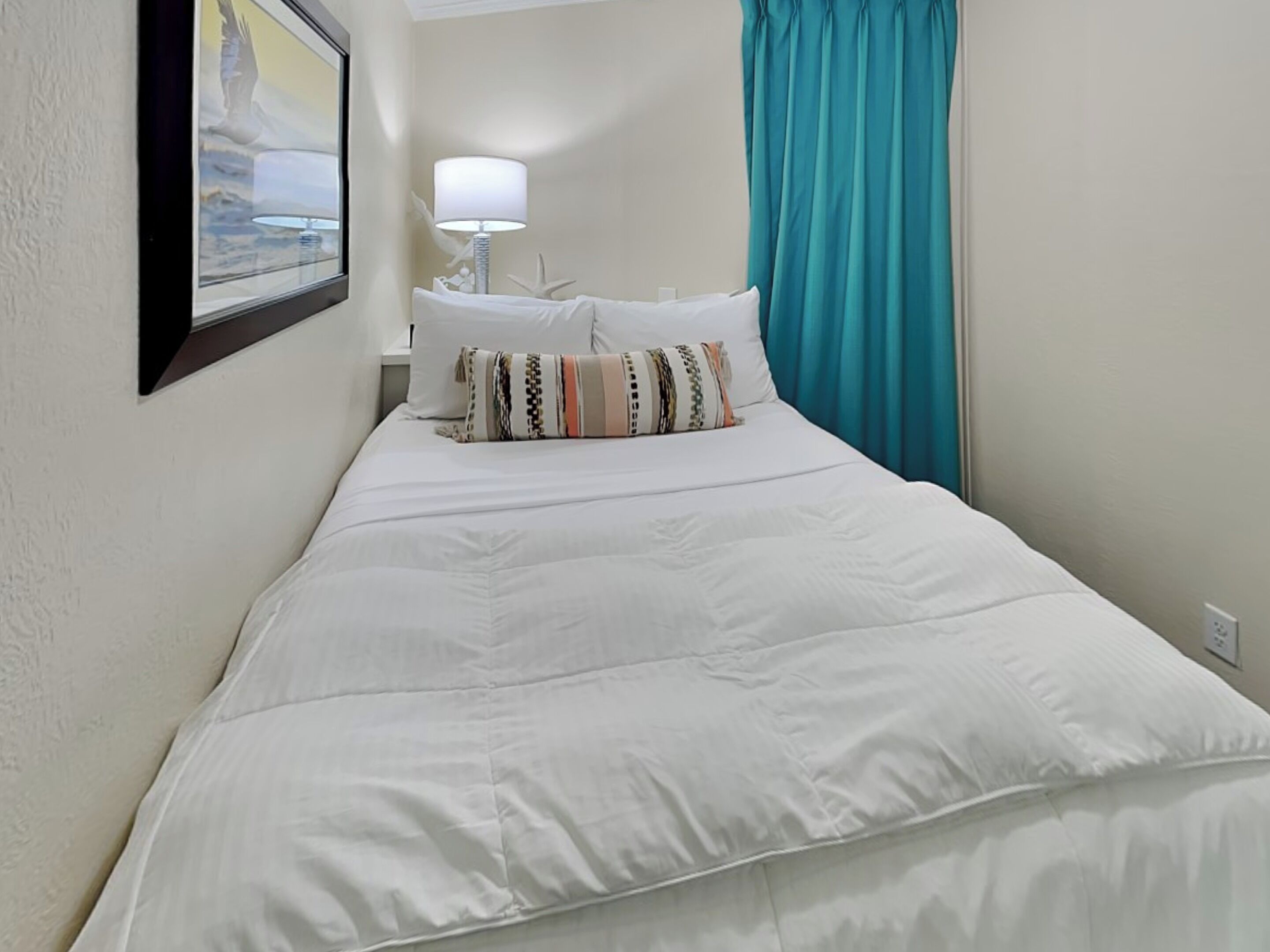 Een opgemaakt bed met witte lakens en meerdere kussens, een witte lamp op een nachtkastje, en een blauwgroene gordijn voor een raam.