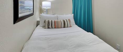 Een opgemaakt bed met witte lakens en meerdere kussens, een witte lamp op een nachtkastje, en een blauwgroene gordijn voor een raam.