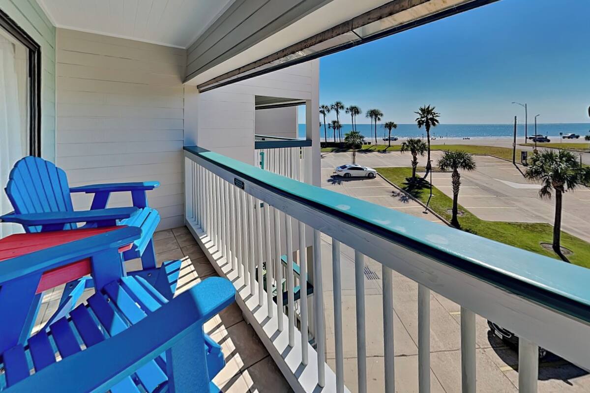 A balcony with blue Adirondack chairs, a view of the beach, and a parking lot with palm trees.