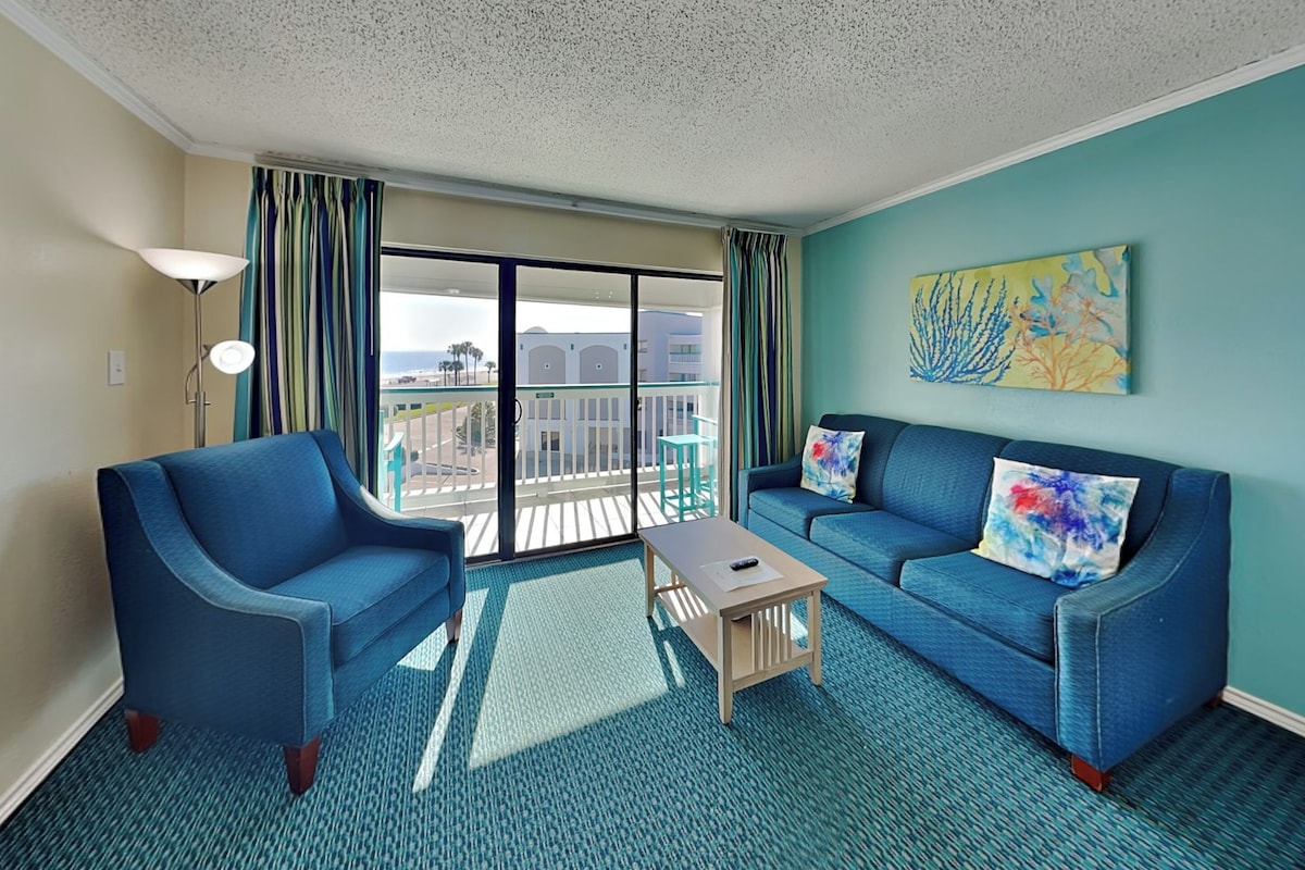 A room with two blue armchairs, a small wooden coffee table, and a blue sofa with decorative pillows. A large window with a view of a building and a balcony is visible.